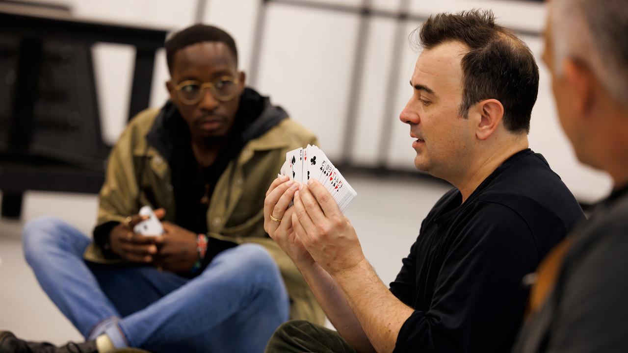Steve Cuiffo is in a navy shirt holding and demonstrating using a deck of cards. A student, seated on the floor, observes intently. The setting is informal and educational, suggestive of a hands-on learning experience.